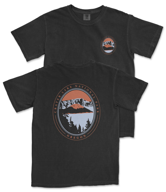 a black t-shirt with a graphic design featuring a mountain landscape and a lake. The design includes a circular emblem with text and an illustration of a mountain and a lake.