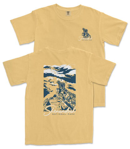 a yellow t-shirt with a graphic design depicting a mountain landscape with a person standing on a rock.