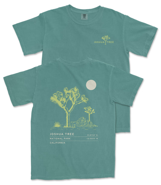 a green t-shirt with a graphic design depicting a desert landscape with a tree and a moon. The t-shirt has text printed on it that reads "JOSUA TREE" and "33.74° N. 115.907 W. CA. N.C. U.S.A."