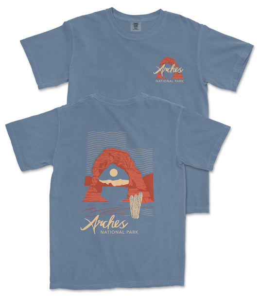 a blue t-shirt with a graphic design depicting a desert landscape with a red rock formation and a cactus. The text on the t-shirt reads "Arches National Park".