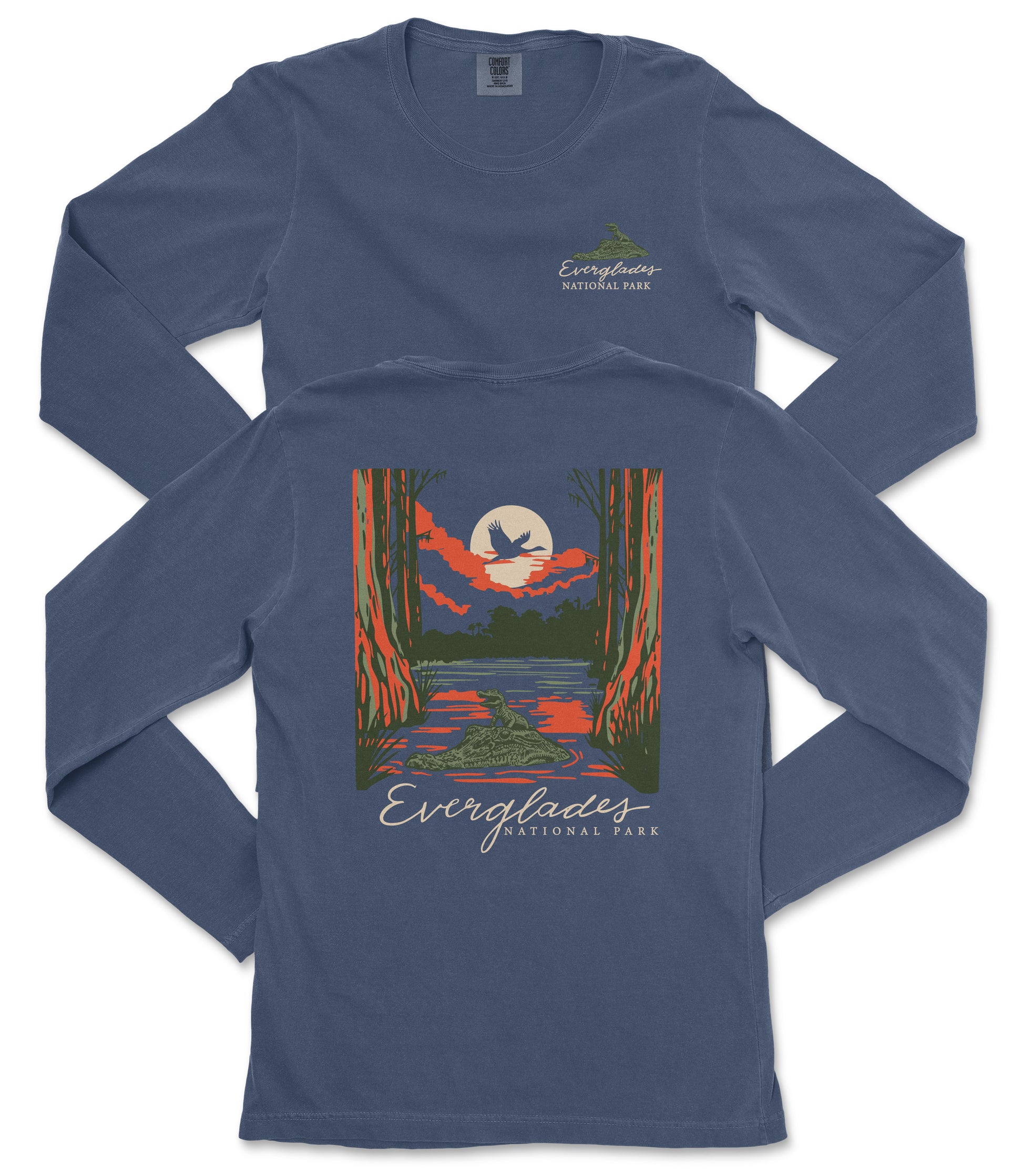 a long-sleeved blue t-shirt with a graphic design depicting a serene landscape with a lake, trees, and a bird flying overhead. The text "EVERGLADES NATIONAL PARK" is printed on the back of the shirt, indicating the design is inspired by Everglades National Park.