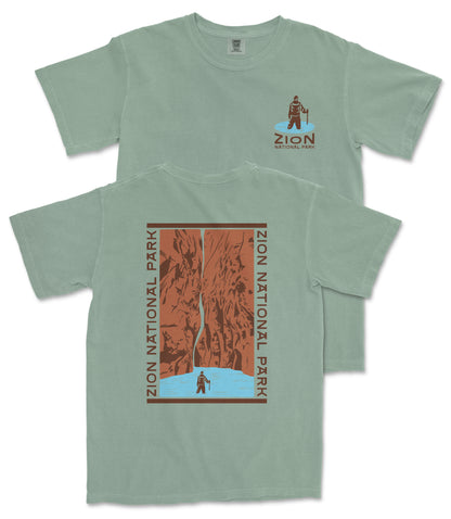 A green t-shirt with a graphic design depicting a person standing in front of a waterfall, surrounded by the text "ZION NATIONAL PARK".