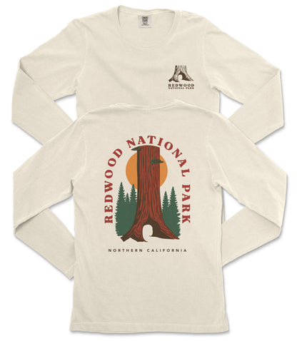 a white long-sleeved t-shirt with a graphic design depicting a tall tree with a sun-like symbol in the center, surrounded by pine trees. The design also includes the text "REDWOOD NATIONAL PARK NORTHERN CALIFORNIA".