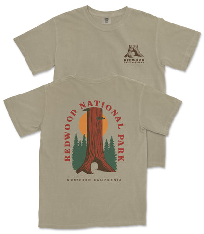 a beige t-shirt with a graphic design depicting a tall tree with a sun-like symbol in the center, surrounded by pine trees. The text on the t-shirt reads "REDWOOD NATIONAL PARK NORTHERN CALIFORNIA".