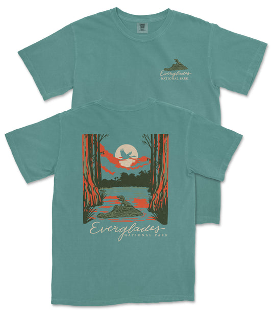 A teal-colored t-shirt with a graphic design depicting a sunset over a forest, with a bird flying in the sky. The text "EVERGLADES NATIONAL PARK" is printed on the back of the t-shirt.