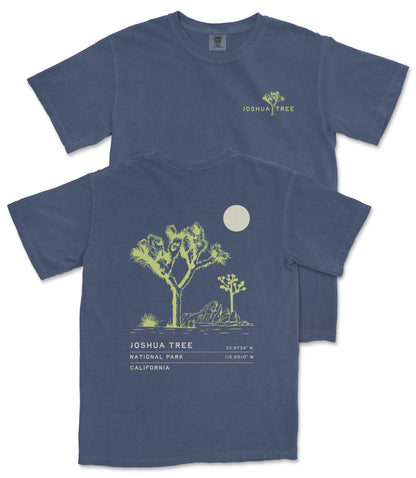 a blue t-shirt with a graphic design depicting a desert landscape with a tree and a moon. The design includes text that reads "JOSUA TREE" and "33.734 N. 115.918 W", indicating the location of Joshua Tree National Park in California.